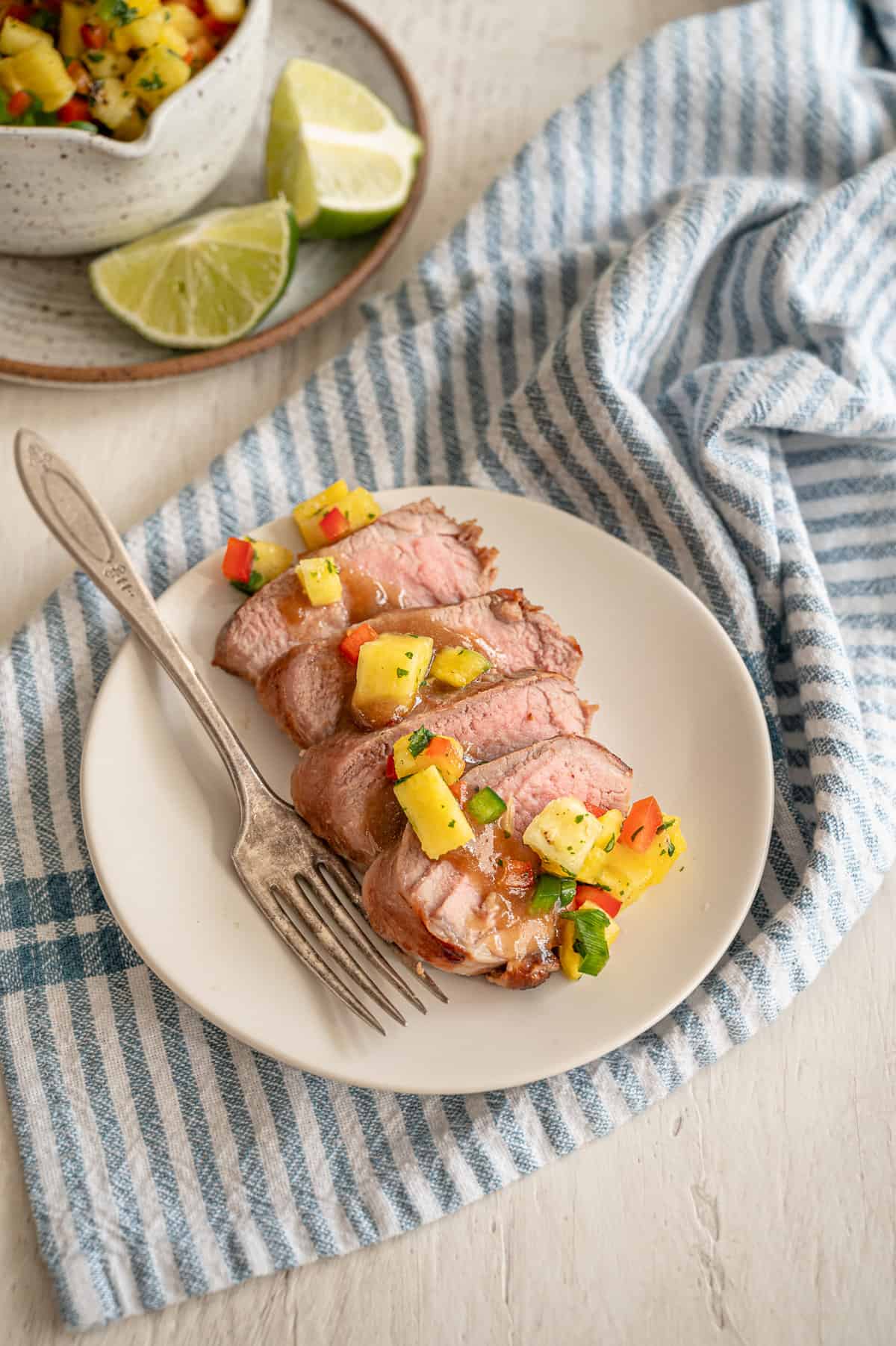 Slices of grilled pork tenderloin on a white plate with pineapple salsa and sauce drizzle on top.