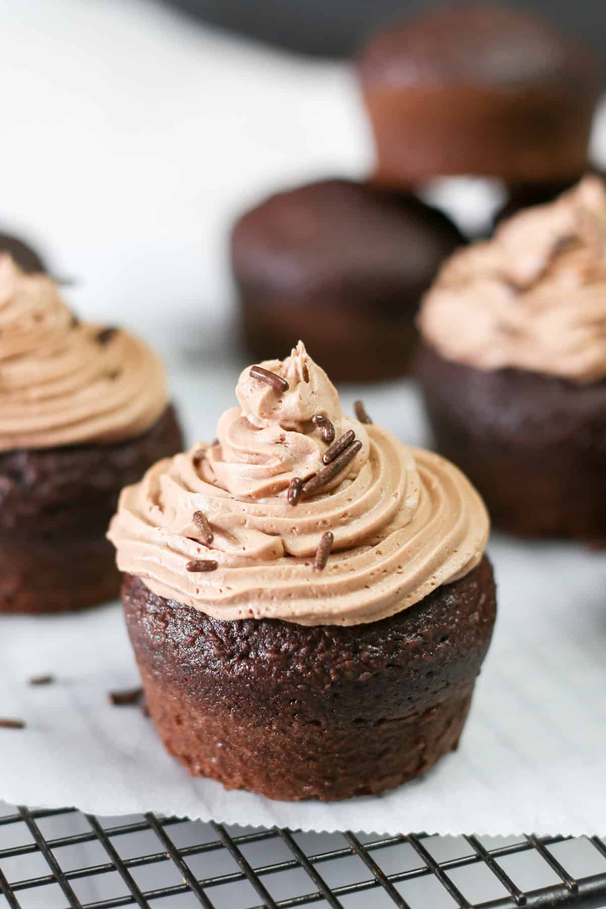 A homemade chocolate cupcake with chocolate buttercream frosting and chocolate sprinkles on a cooling rack.