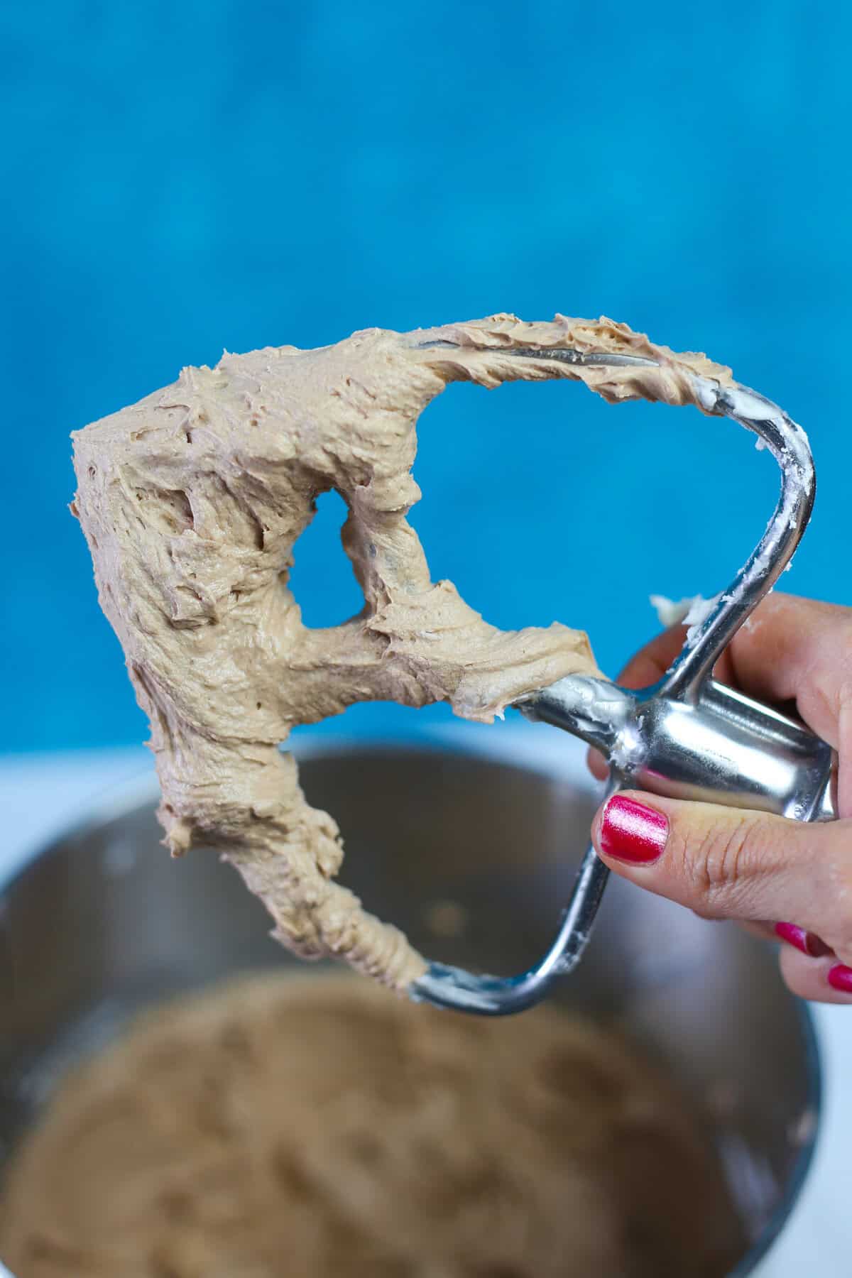 A paddle attachment from a mixer covered in chocolate buttercream frosting being held above the mixing bowl full of frosting.