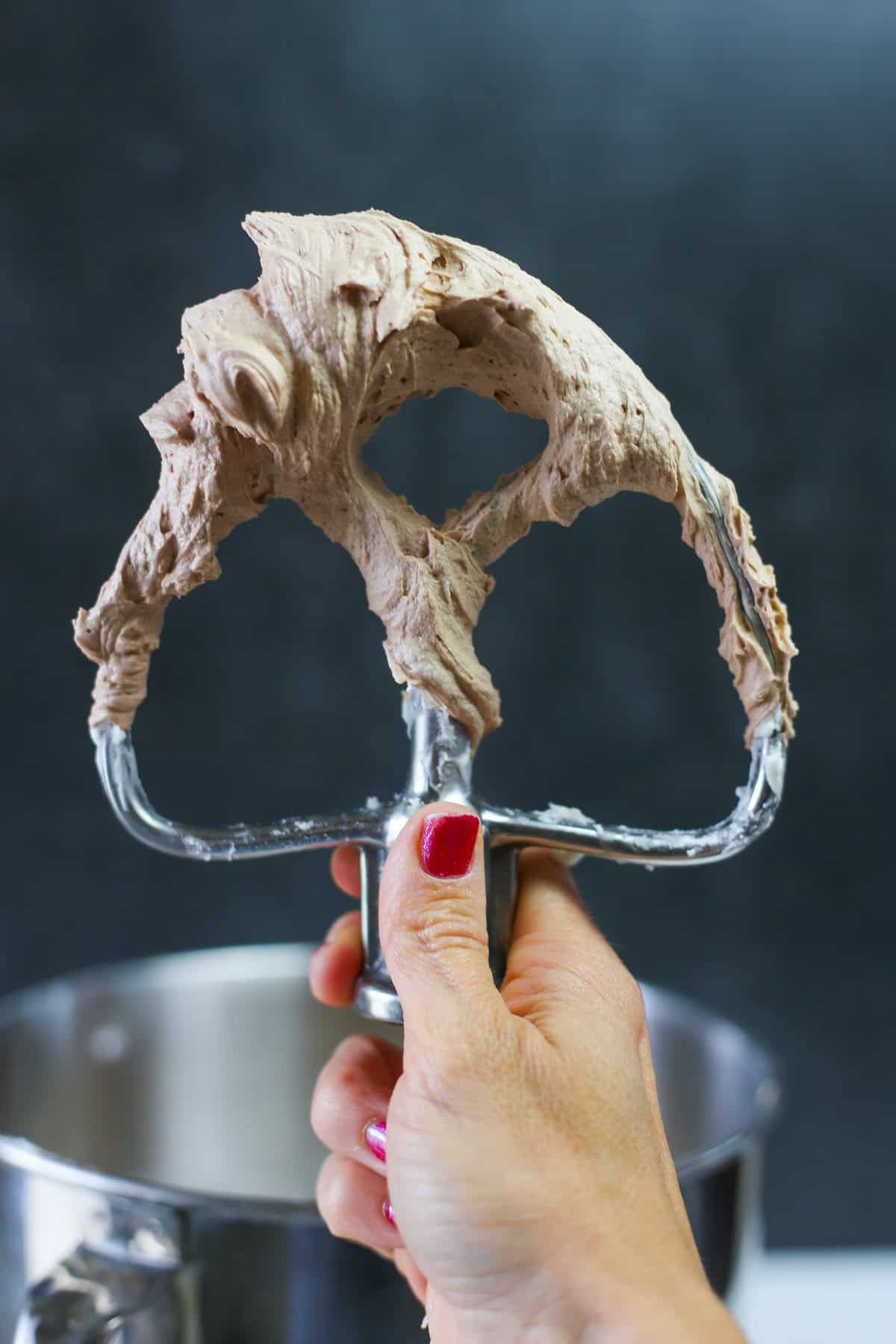 A hand holding up a mixer paddle attachment covered in chocolate buttercream frosting.