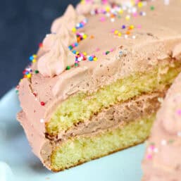 A double-layer yellow cake with chocolate buttercream frosting on a cake stand with a piece missing.