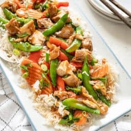 A platter of honey bourbon chicken with rice and stir-fried veggies with chopsticks and a stack of plates.