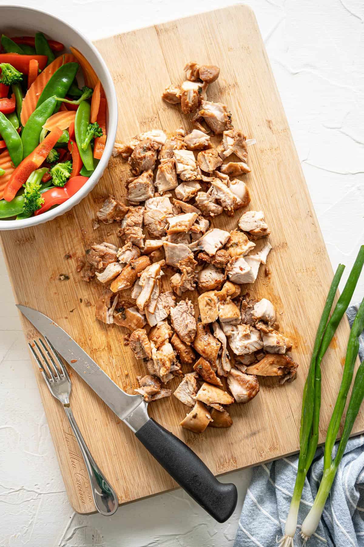 Diced chicken on a wooden cutting board with a bowl of stir-fry veggies.