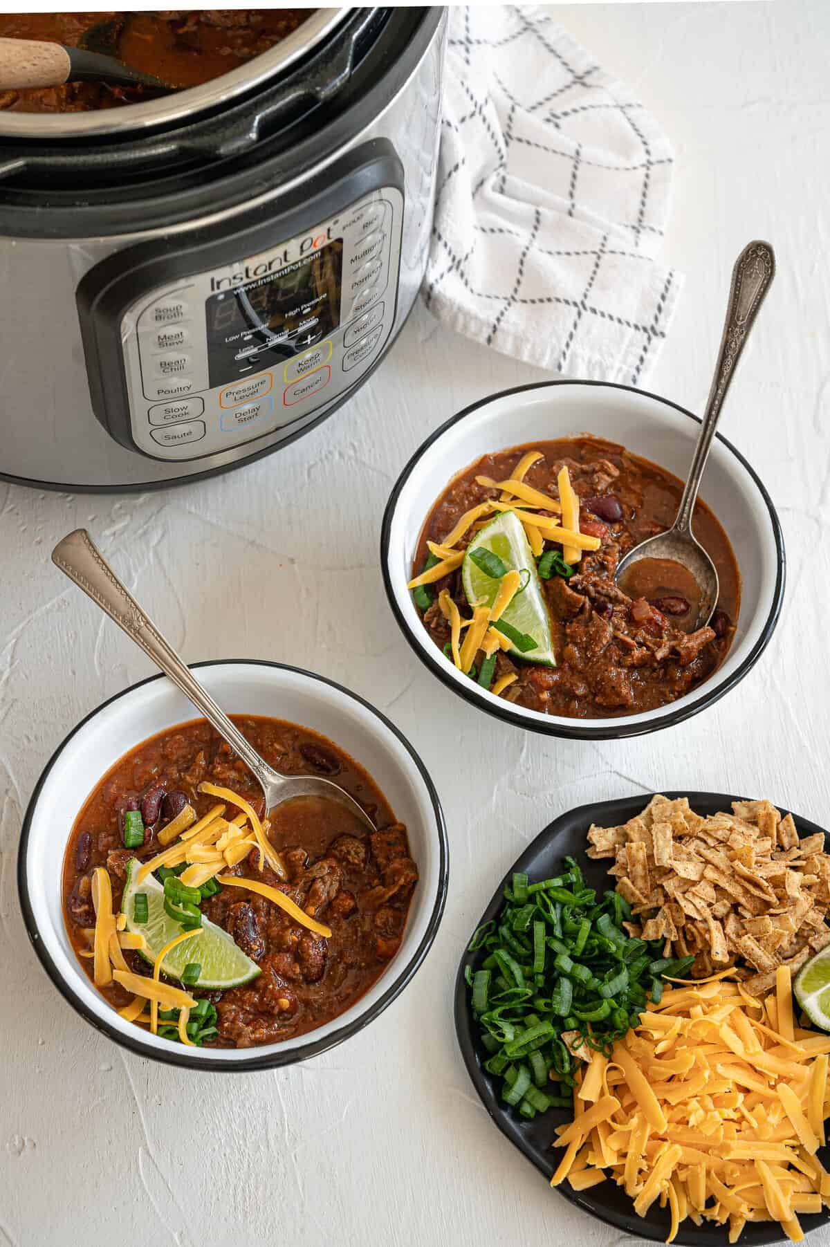 Steak chili served in two bowls with a place of toppings including chopped chives, lime wedges, tortilla strips, and shredded cheddar cheese.