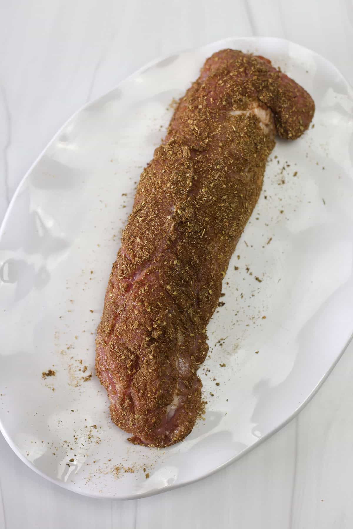 Pork tenderloin on a white platter covered with an herb rub ready to be cooked.