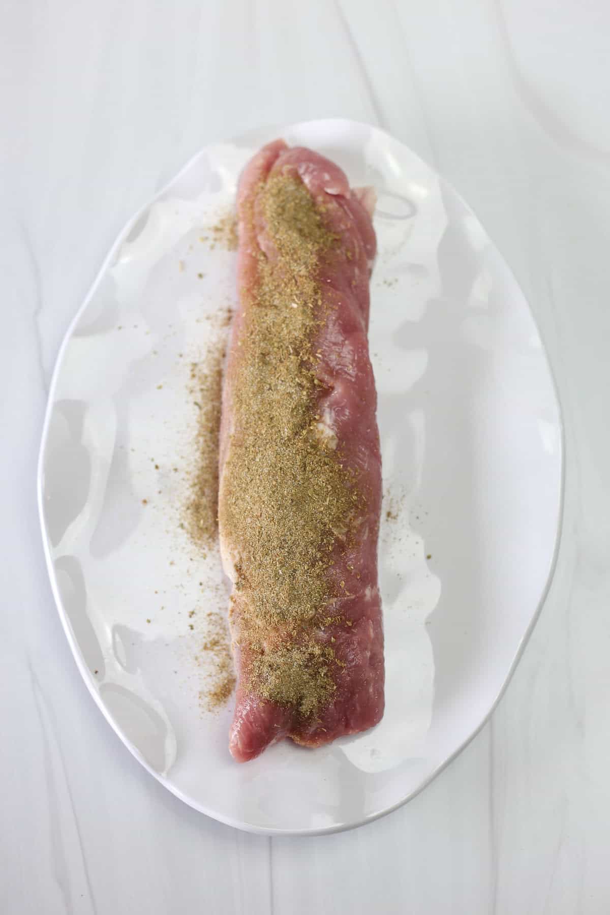 A pork tenderloin with a dry rub sprinkled over top ready to be rubbed in.