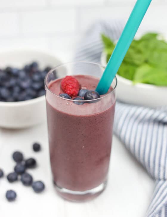 A purple smoothie in a glass cup with berries on top and in the background.