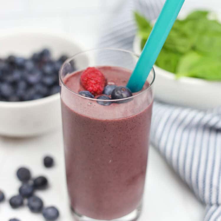 A purple smoothie in a glass cup with berries on top and in the background.