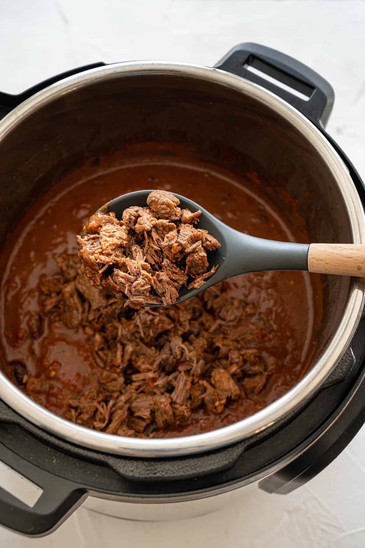 Wooden spoon above an Instant Pot holding shredded beef stew meat to stir into the rest of the Steak Chili ingredients.