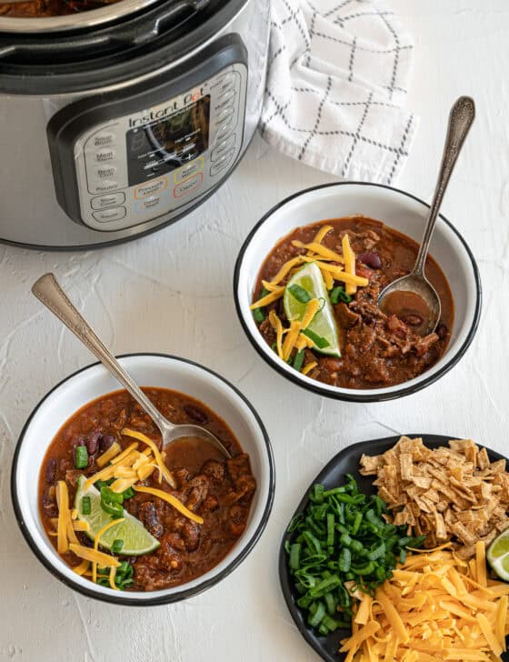 Two bowls of steak chili in front of an Instant Pot with a lime wedge, chopped green onions, and shredded cheese on top of each.