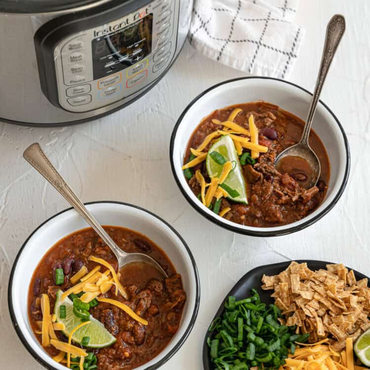 Two bowls of steak chili in front of an Instant Pot with a lime wedge, chopped green onions, and shredded cheese on top of each.