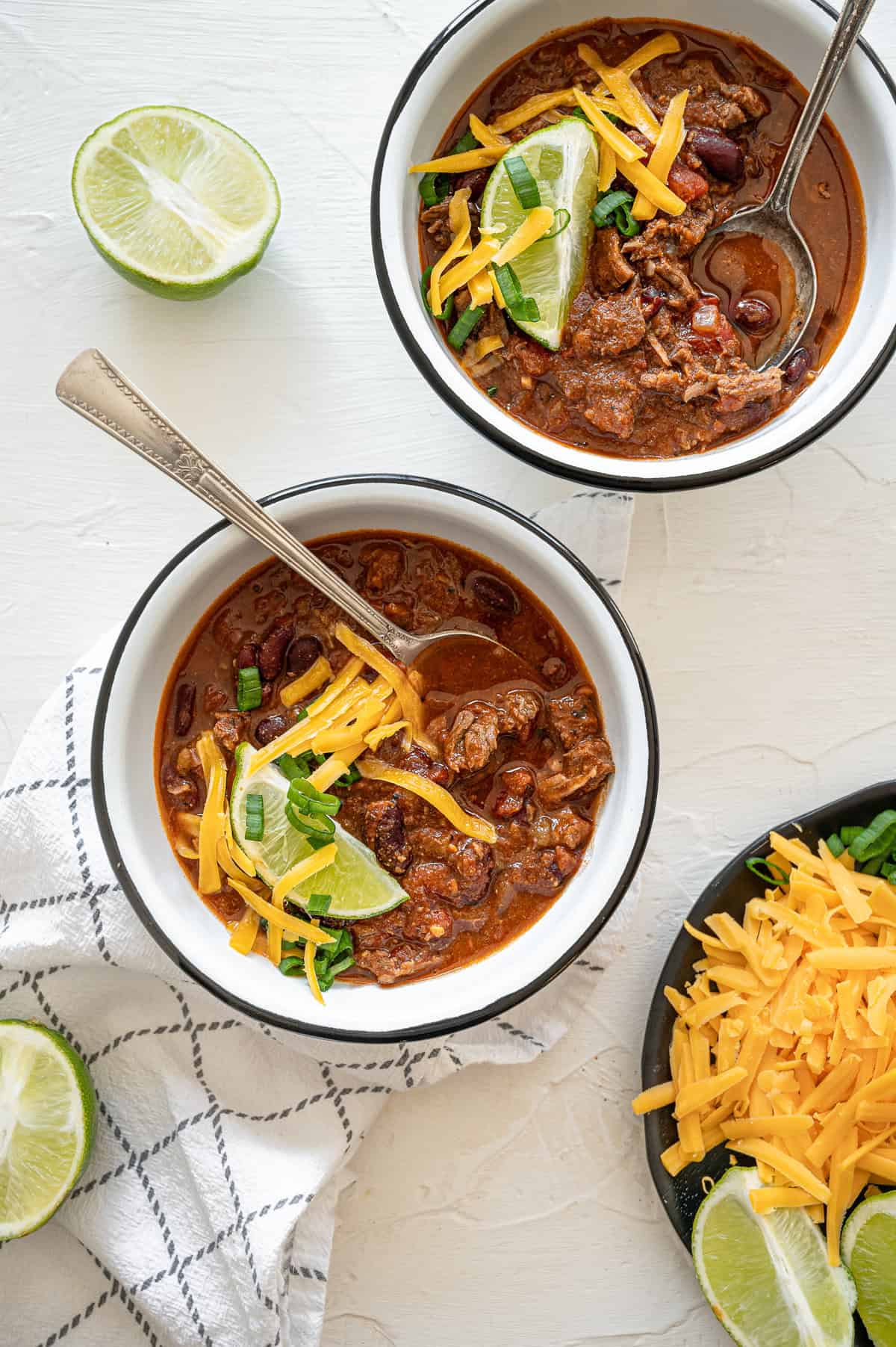 Two bowls of steak chili with a lime wedge, shredded cheddar cheese, and chopped green onions on top of each.