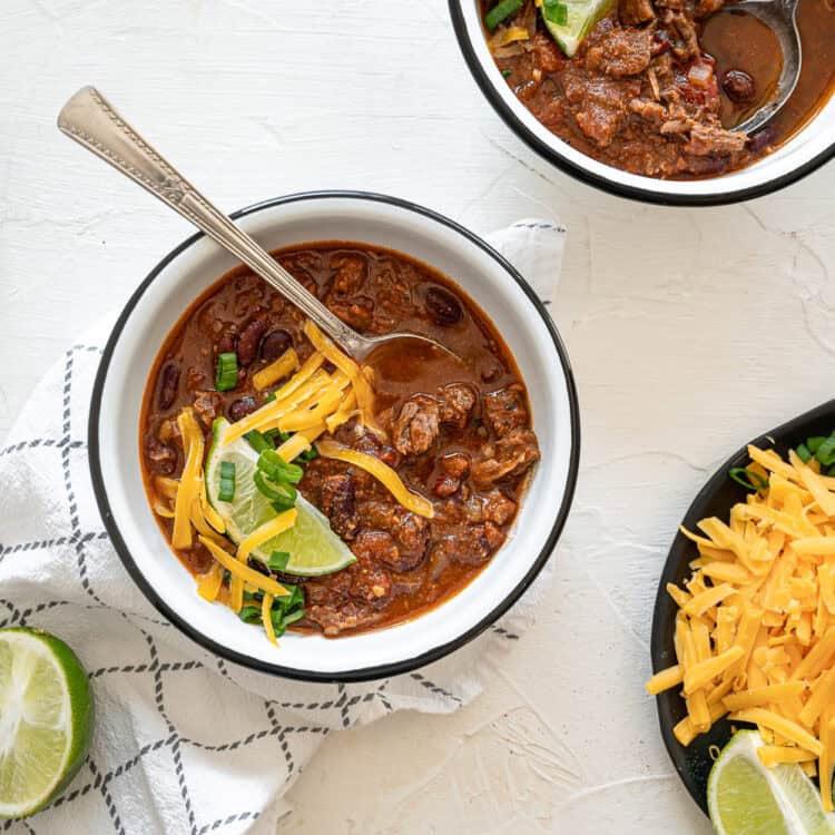 A bowl of steak chili with a plate of toppings including shredded cheese, chopped green onions, tortilla strips, and lime wedges sitting next to it.