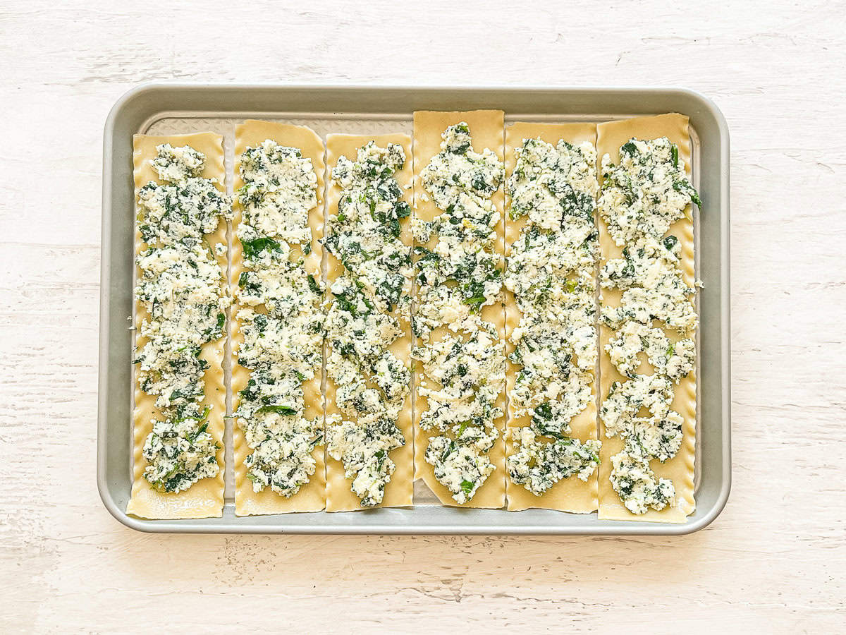 Cooked lasagna noodles on a baking sheet lined up with a mixture of cheeses, herbs, and spinach spread on each.