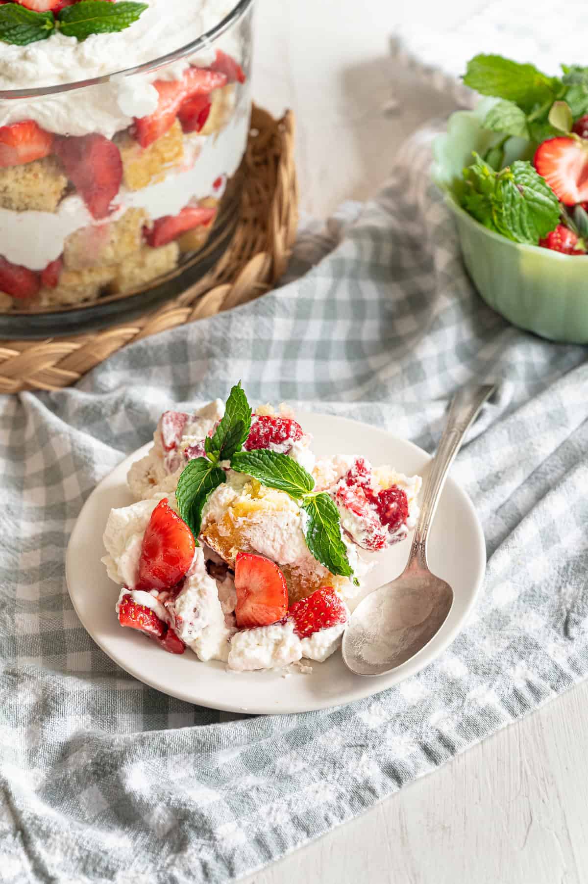 A serving of strawberry shortcake trifle on a white plate with the trifle dish behind it.