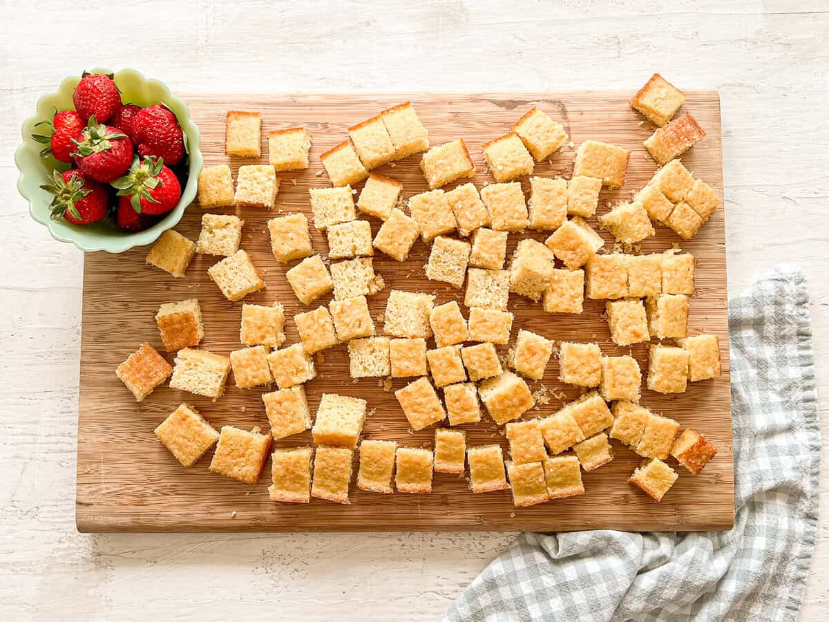 A wooden cutting board with cubed pieces of yellow cake and a small bowl of strawberries on it.