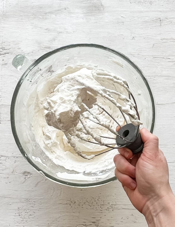 A hand holding a beater from a mixer with fresh whipped cream on it.