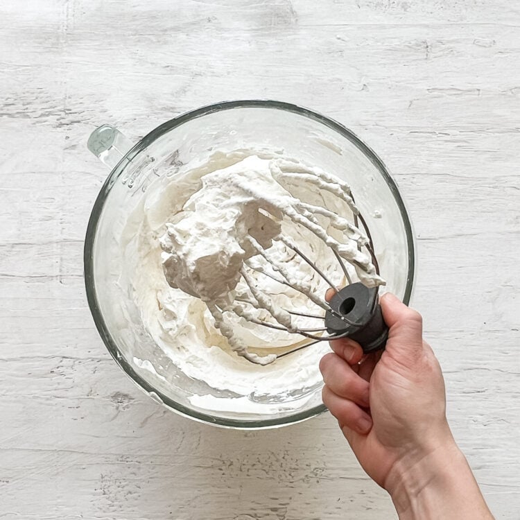 A hand holding a beater from a mixer with fresh whipped cream on it.