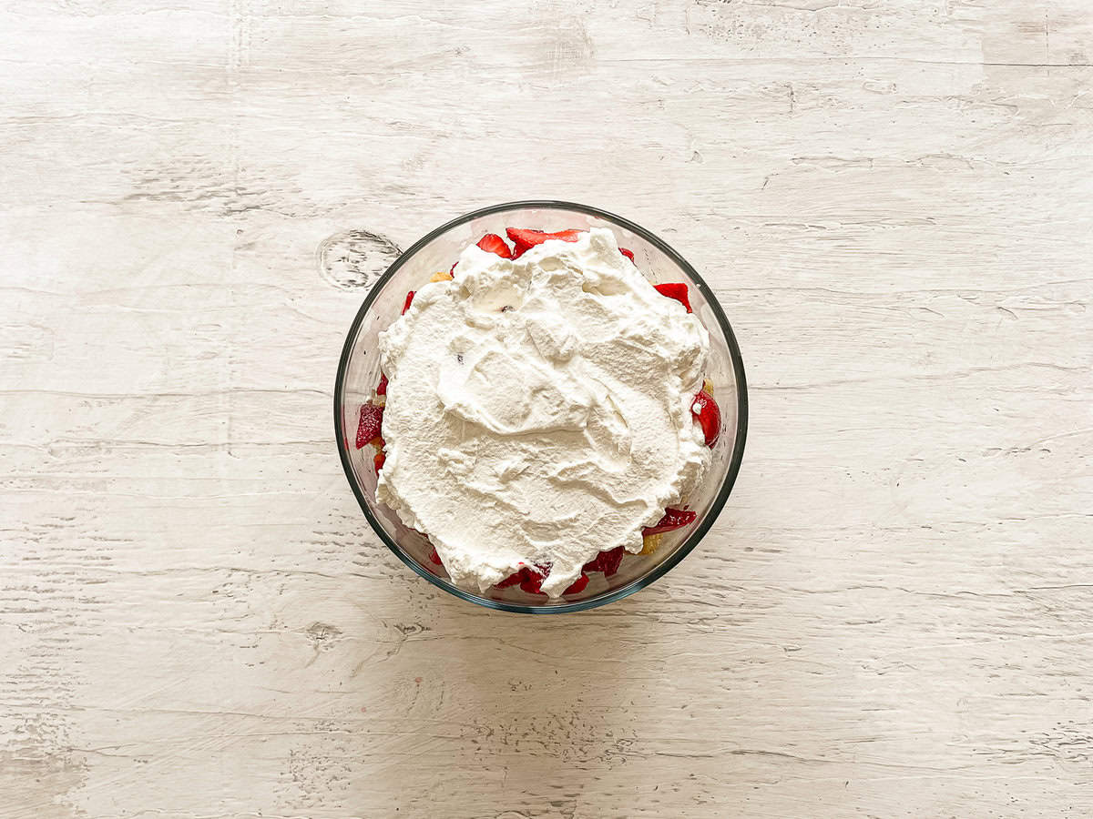 A glass dish with layers yellow cake cubes, then sliced strawberries, and finally fresh whipped cream spread on top.