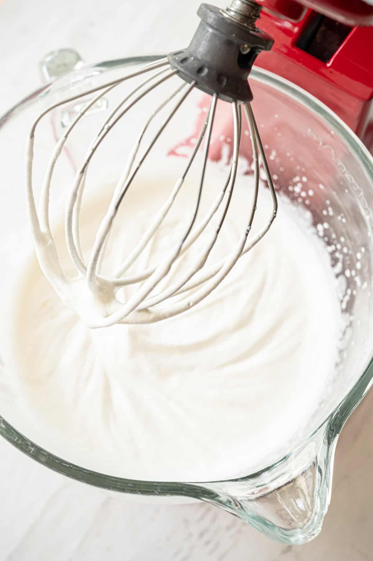 The beater of a mixer above a glass mixing bowl with fresh whipped cream.