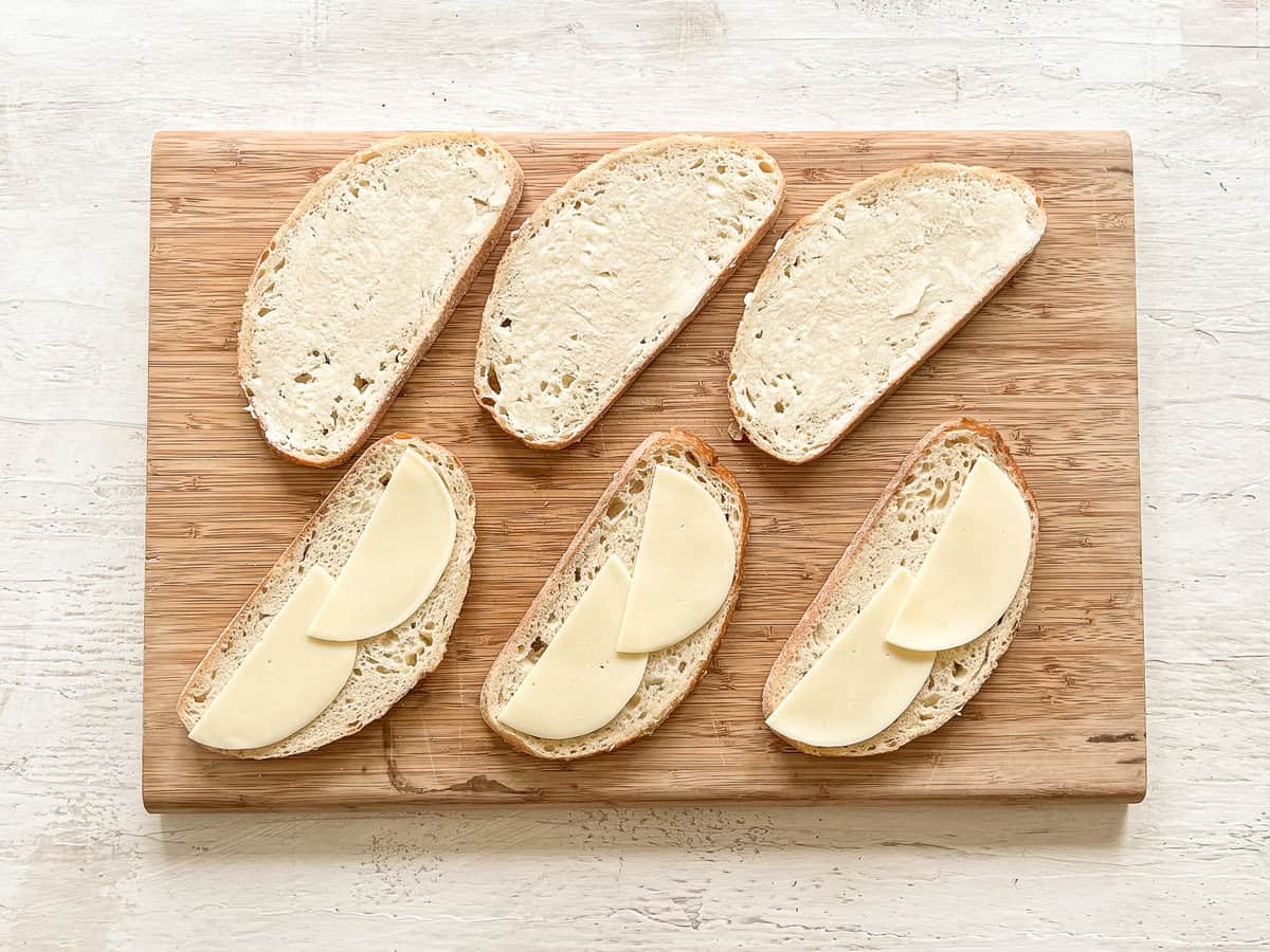 Slices of bread lined up on a cutting board, half with butter, and the other half with provolone slices on them.