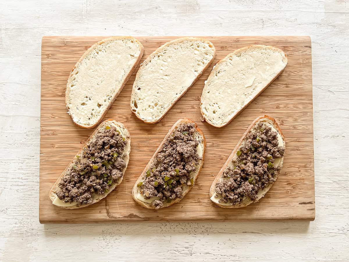 Slices of bread lined up on a cutting board, half of them with provolone slices and a ground beef mixture to make ground beef Philly cheesesteaks.
