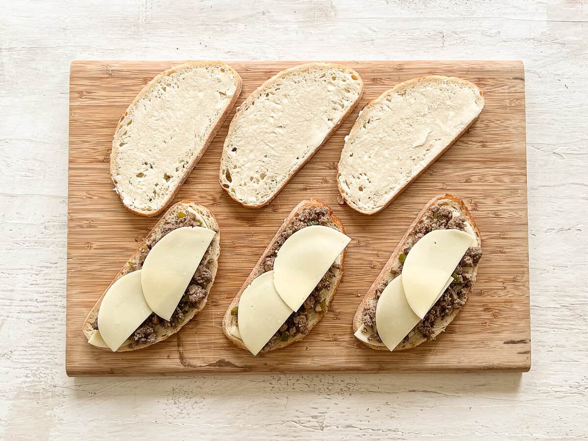 Slices of bread lined up on a cutting board, half of them with provolone cheese, ground beef mixture and more provolone cheese on top ready to be toasted ground beef Philly cheesesteaks.