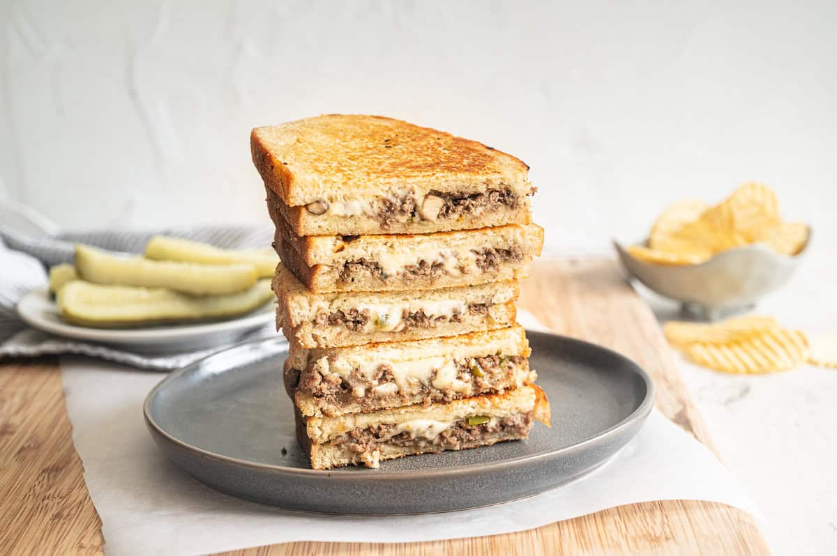 Ground beef Philly cheesesteaks cut in half and served on a plate with pickle spears and chips.