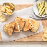 Ground beef Philly cheesesteaks cut in half and lined up on a wooden cutting board with bowls of chips and pickles on the side.