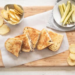 Ground beef Philly cheesesteaks cut in half and lined up on a wooden cutting board with bowls of chips and pickles on the side.