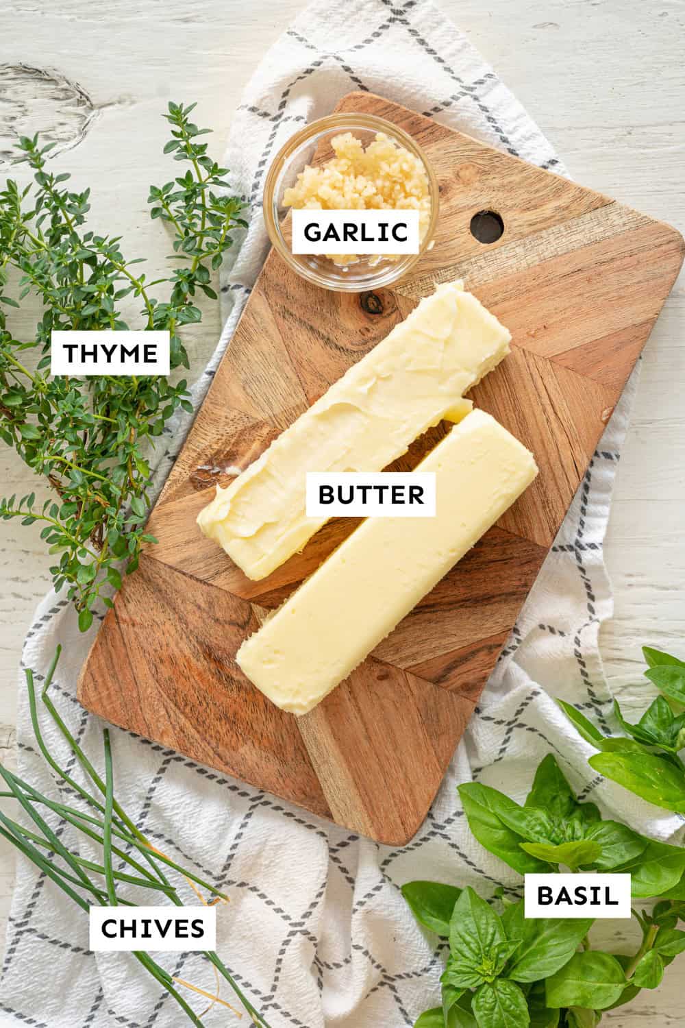 Two softened butter sticks on a cutting board with a small bowl of minced garlic and fresh herbs around it.