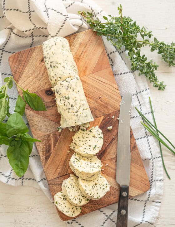 Log of garlic herb butter on a wooden cutting board with part of it already in slices for serving.