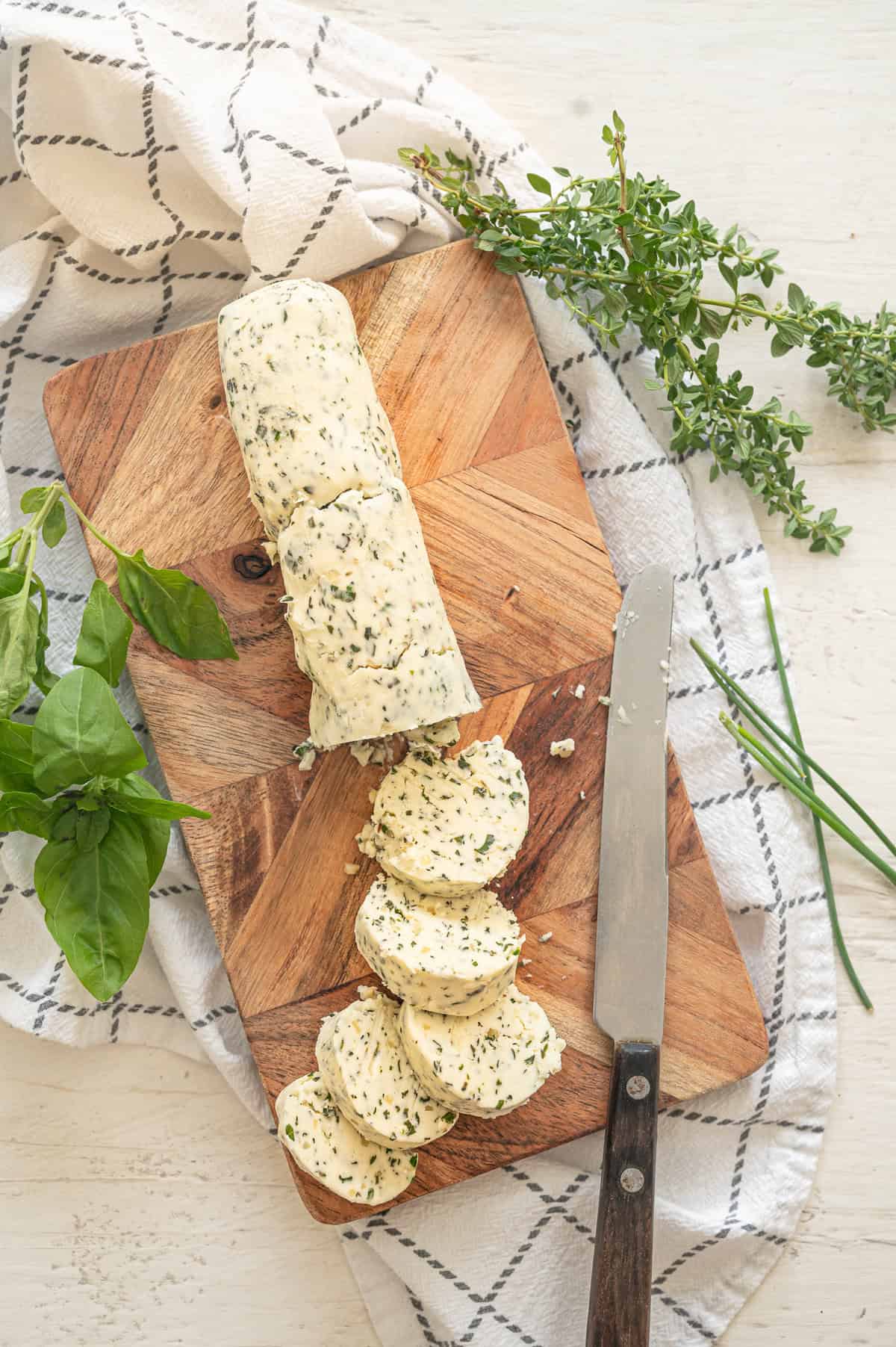 Log of garlic herb butter on a wooden cutting board with part of it already in slices for serving.