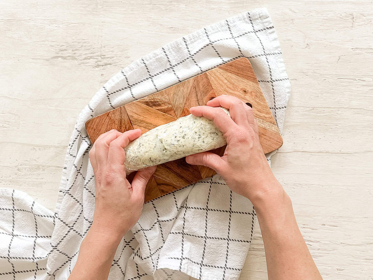 Two hands forming a roll of garlic herb butter in plastic wrap on a wooden cutting board.