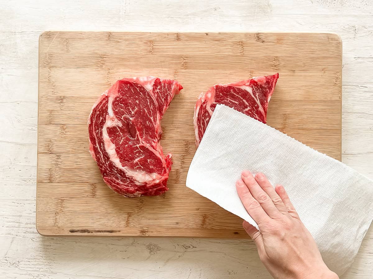 A hand using a paper towel to dry two steaks on a cutting board.