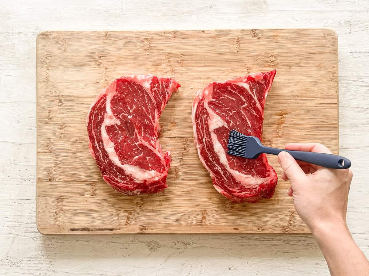 A hand brushing two steaks on a cutting board with oil.