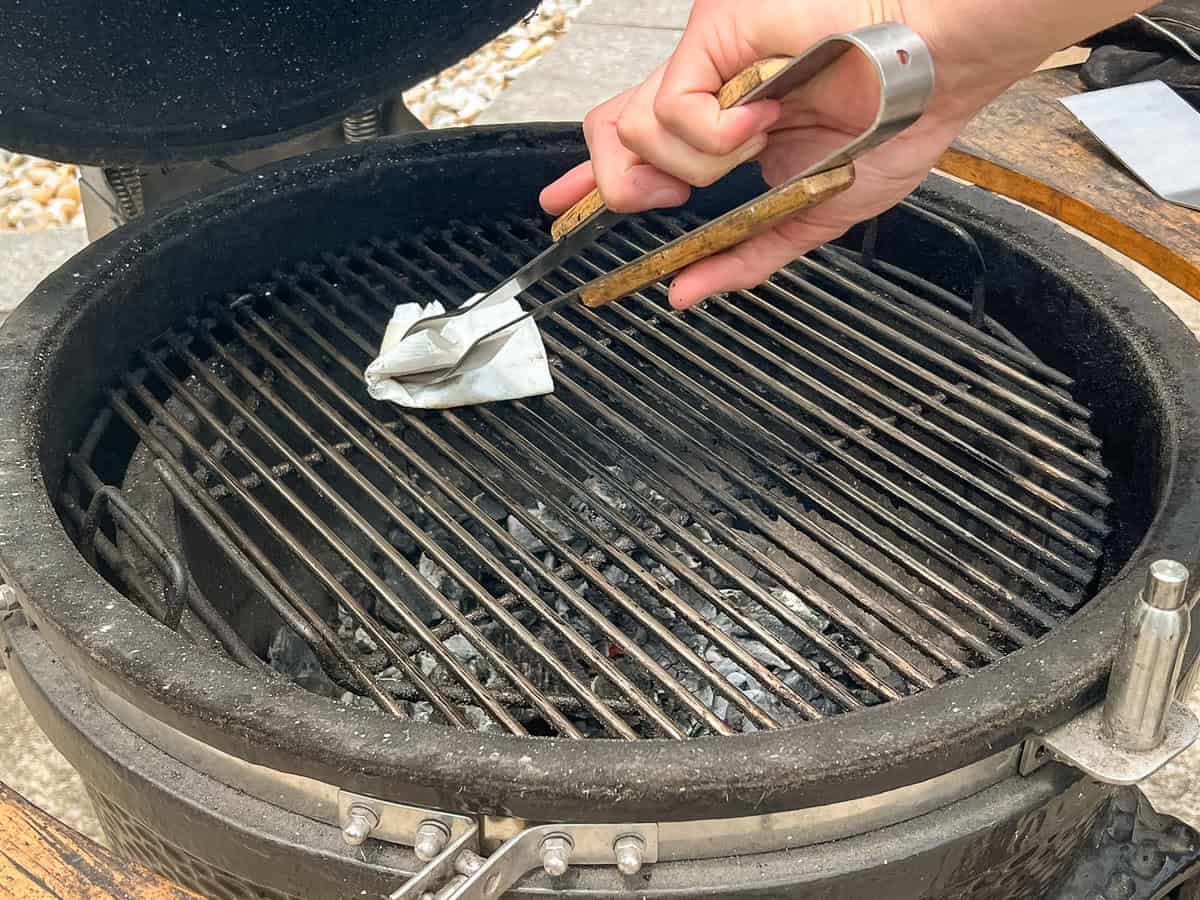 A hand using tongs with an avocado oil-soaked paper towel to oil the grates on a grill.