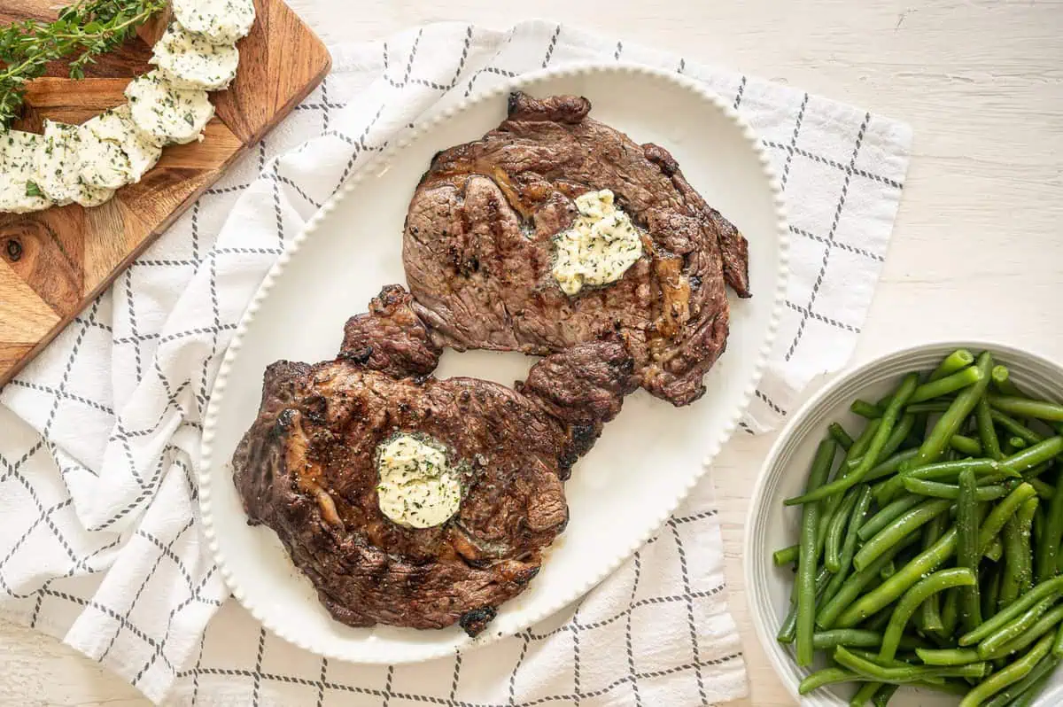 Two steaks with garlic herb butter on a platter and a bowl of green beans next to it.