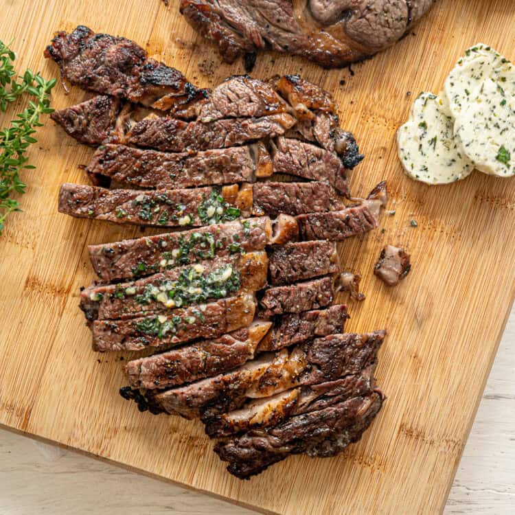 Sliced grilled steak on a wooden cutting board with garlic herb butter slices next to it.