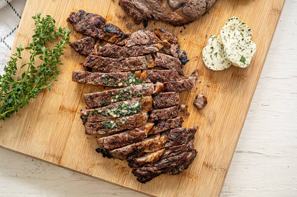 Sliced grilled steak on a wooden cutting board with garlic herb butter slices next to it.