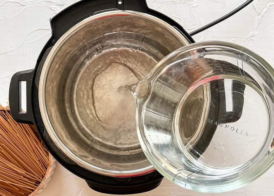 Water being poured into an Instant Pot with a bowl of uncooked whole wheat spaghetti noodles next to it.