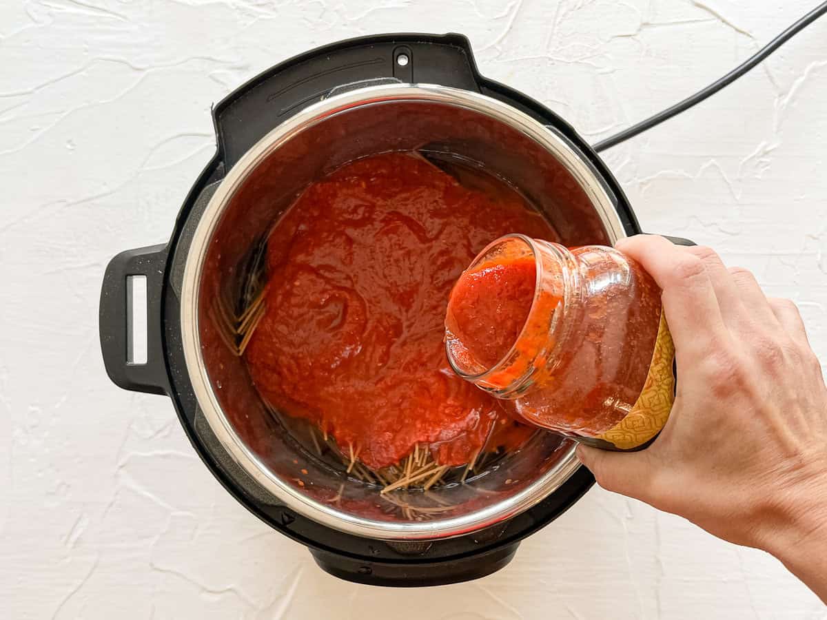 A jar of marinara being poured on top of whole wheat spaghetti noodles in an Instant Pot.