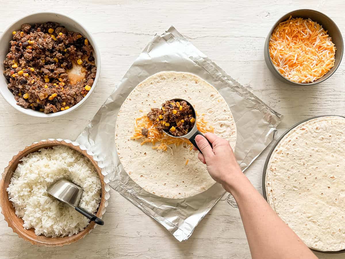 A hand putting a scoop of burrito beef mixture on a tortilla at burrito-making station with bowls of rice, cheese, beef mixture, and tortillas.