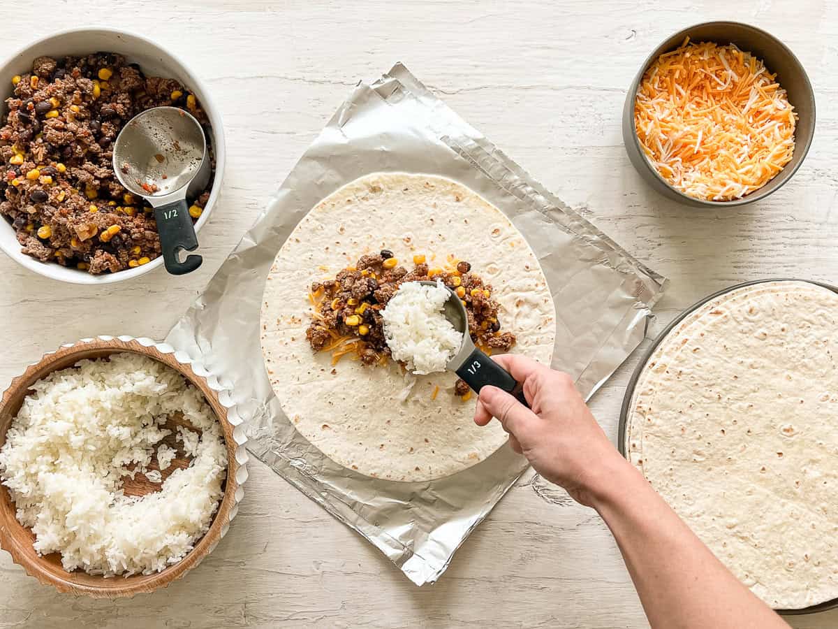 A hand putting a scoop of rice on a tortilla at a burrito-making station with bowls of rice, beef mixture, cheese, and tortillas.