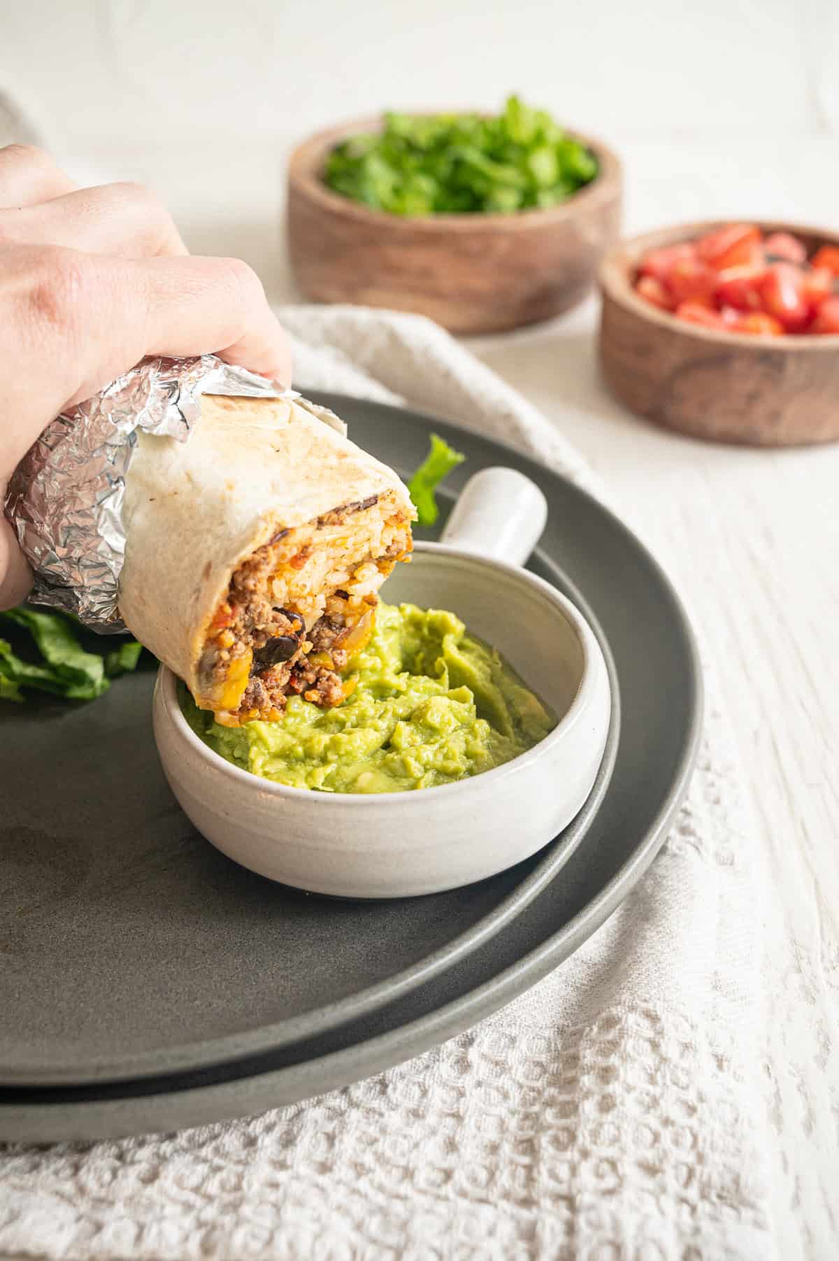 A hand dipping a beef burrito into a bowl of guacamole.