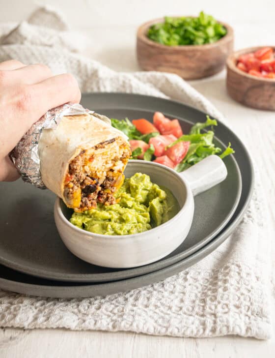 Half of a beef burrito being dipped in a bowl of guacamole.