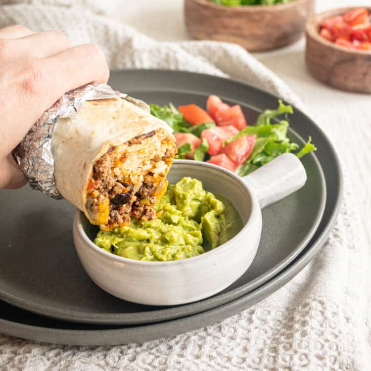 Half of a beef burrito being dipped in a bowl of guacamole.