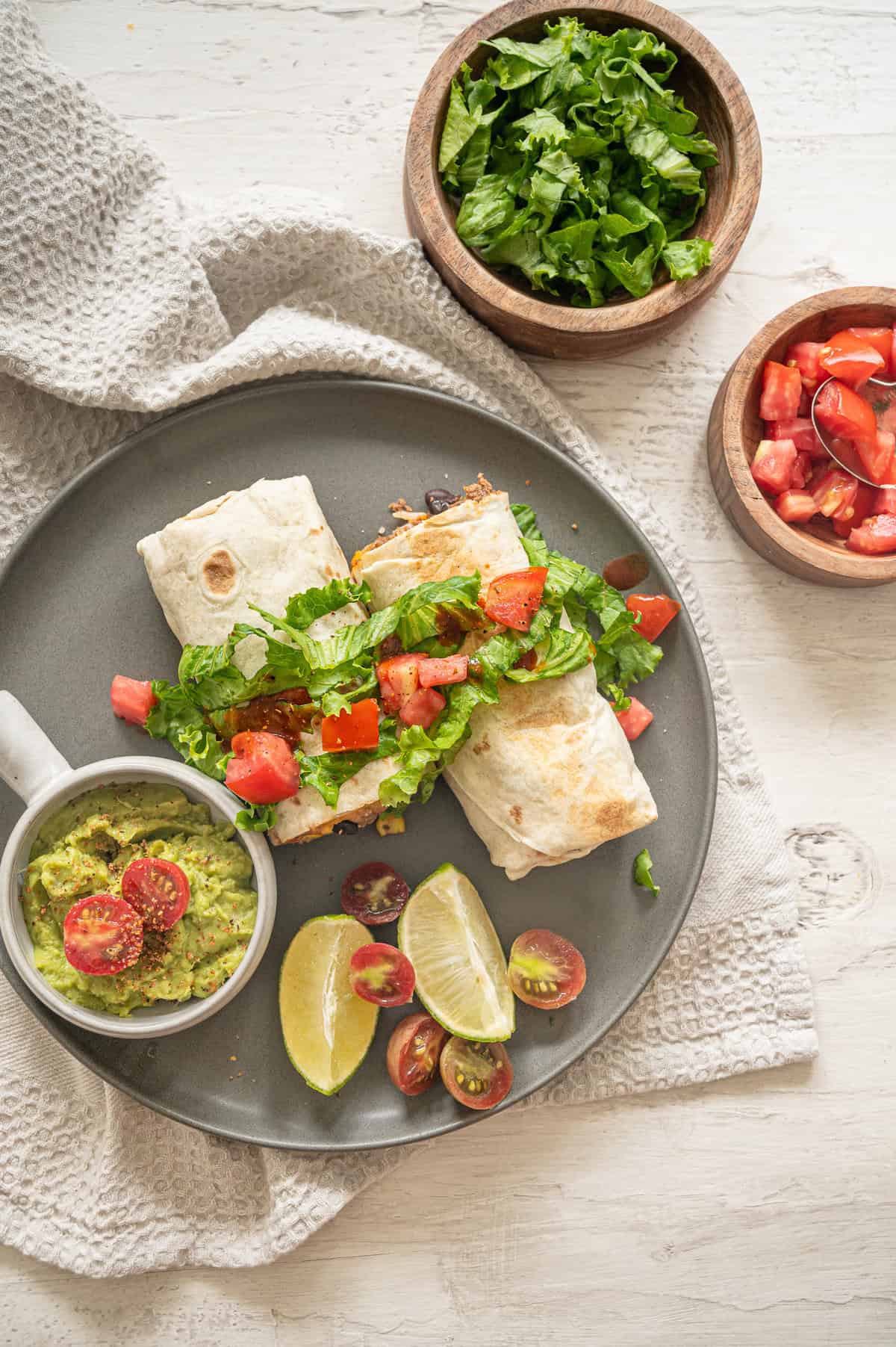 Beef burritos on a plate with shredded lettuce and chopped cherry tomatoes on top and a bowl of guacamole on the side.