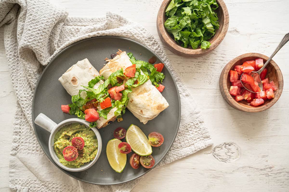 Beef burritos on a plate with a bowls of guacamole, chopped cherry tomatoes, and shredded lettuce.