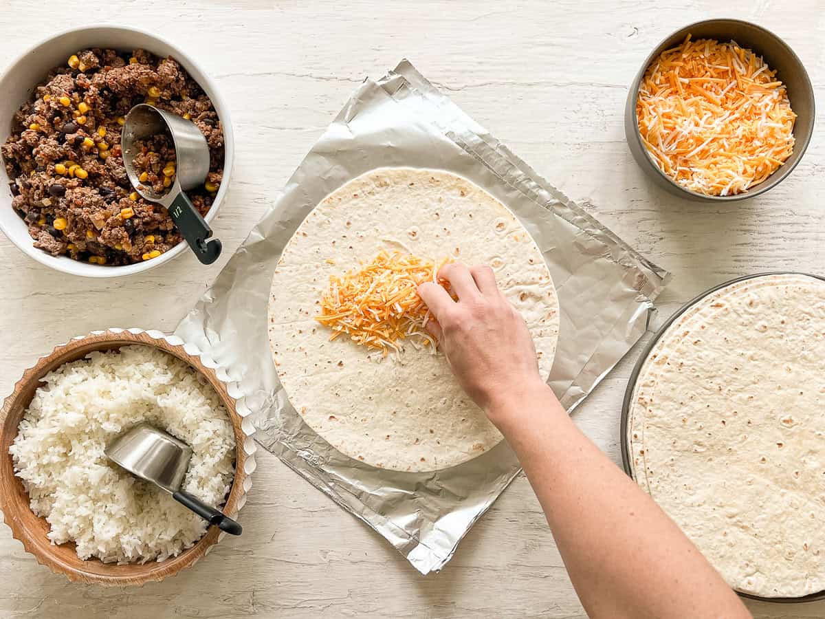 A hand putting cheese on a tortilla at a burrito making station with bowls of beef mixture, rice, tortillas, and cheese.